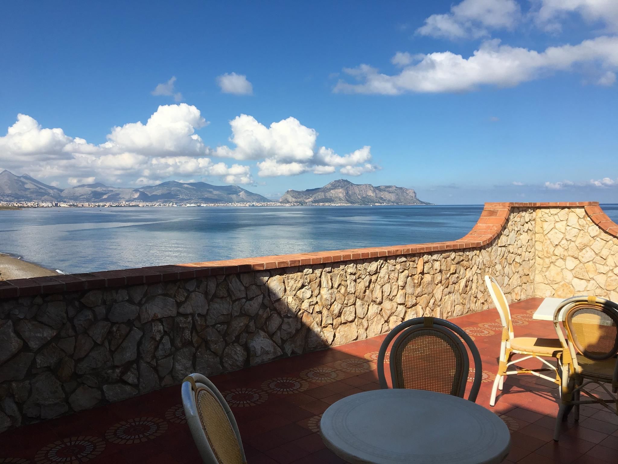 Un muro de piedra da a un mar tranquilo con montañas en la distancia. Hay dos sillas de mimbre y una pequeña mesa redonda en el patio.