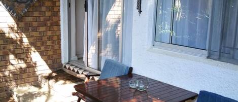 Un patio con una mesa y sillas de madera, un muro de ladrillo y una ventana con cortinas.