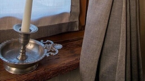 A lit candle on a metal stand next to a window with sheer curtains tied back with a fabric tie. - La cor de Jeremie (Montjovet)