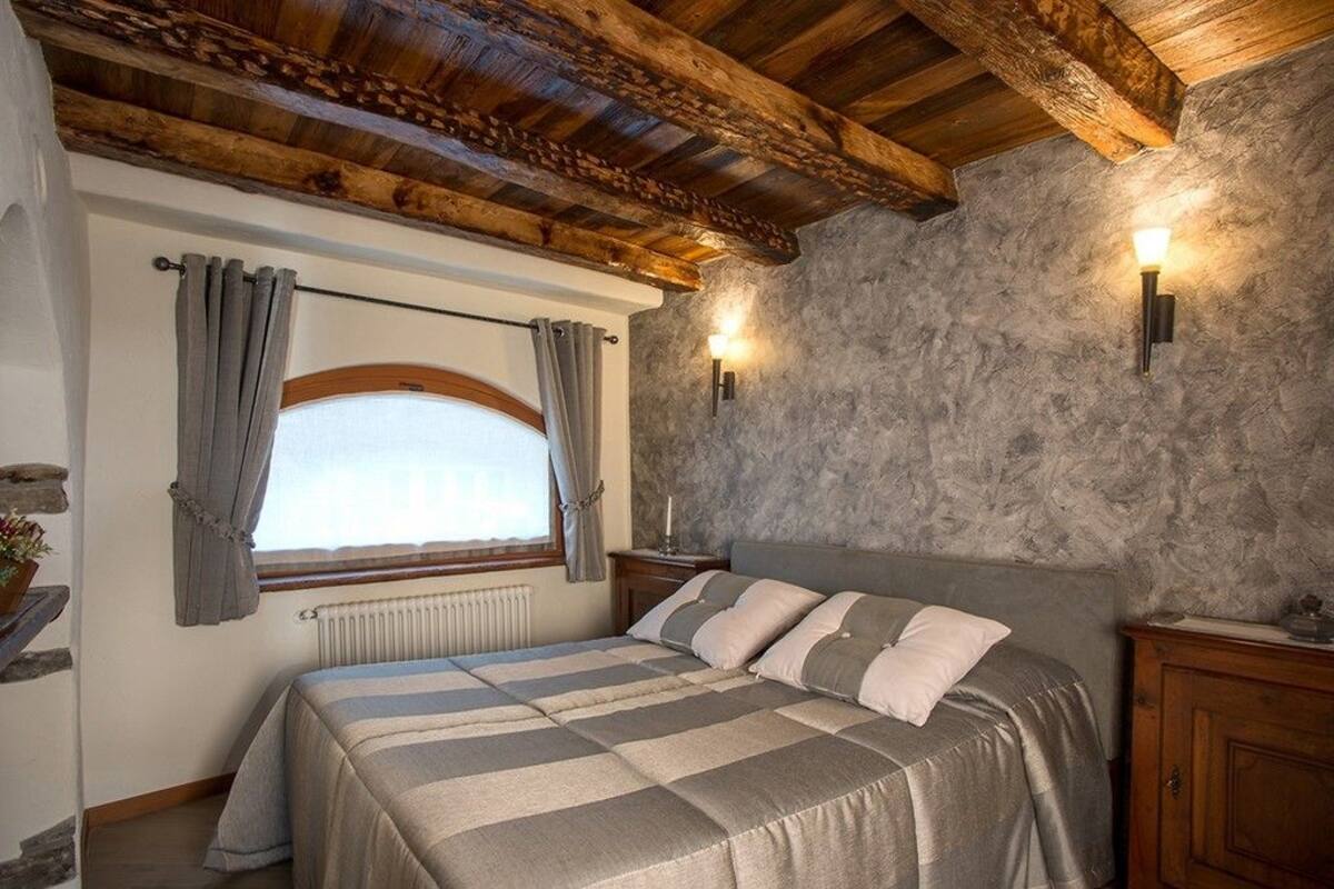 Un dormitorio con cama, vigas de madera en el techo, paredes de piedra, una ventana con cortinas e iluminación empotrada.