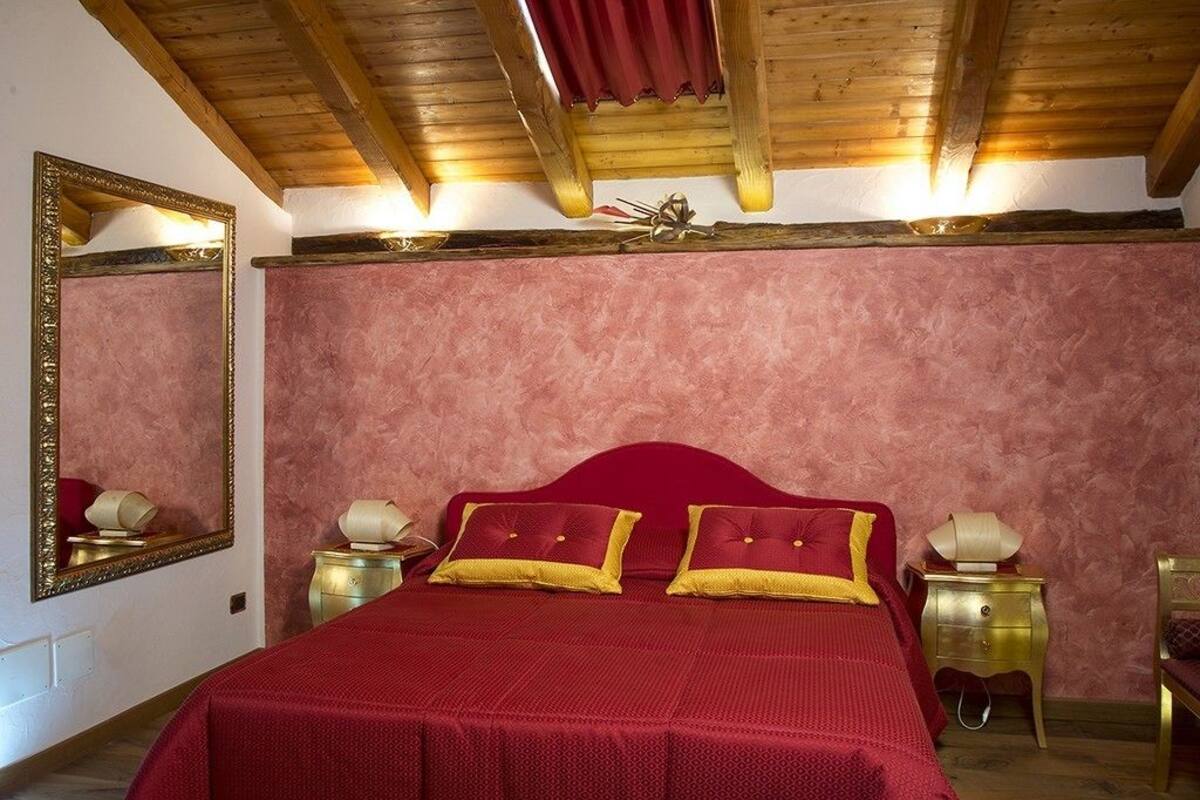 Un dormitorio con colcha roja, dos mesitas de noche con lámparas, un espejo grande y techo de madera.