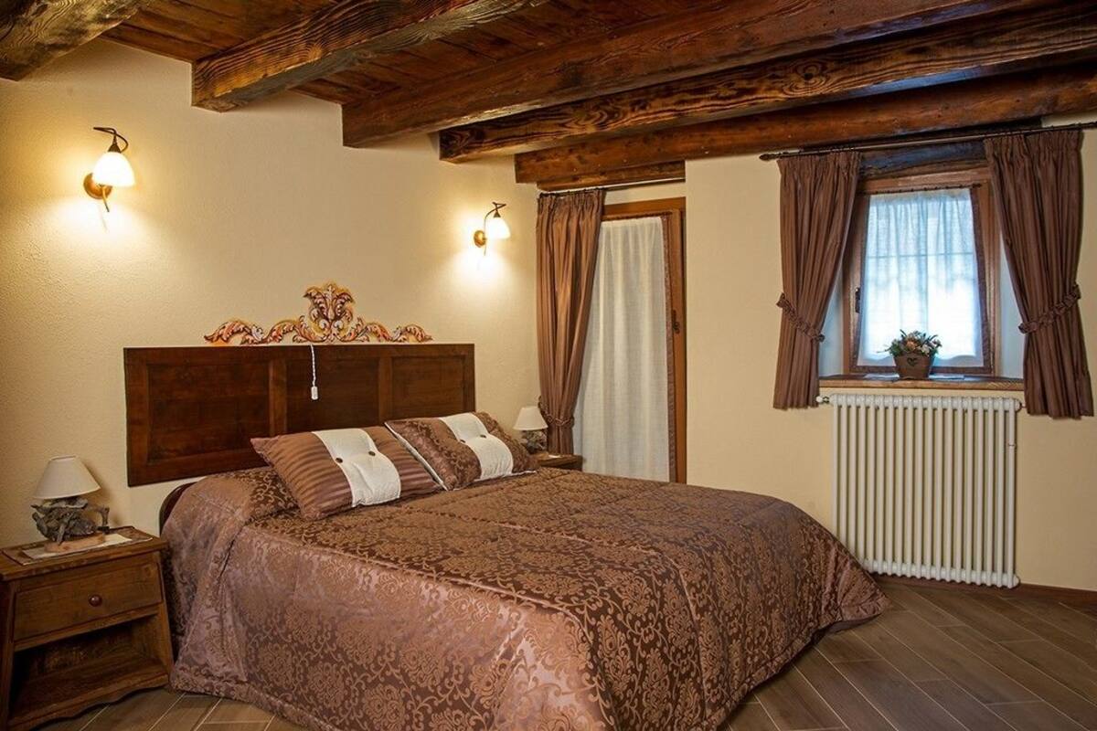 Un dormitorio con cabecera de madera, una cama grande con ropa de cama estampada, dos lámparas de noche, una mesita de noche, una ventana con cortinas y un radiador.