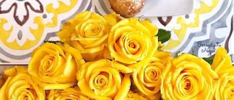 A table with a patterned tablecloth, a bouquet of yellow roses, a small plate with a pastry, and a cup of tea.