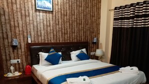 Habitación de hotel con pared de paneles de madera, cama con ropa de cama blanca y detalles azules, mesita de noche con lámpara y ventana con cortinas a rayas negras y blancas.