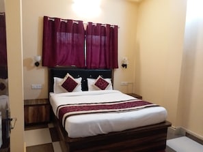 A neatly made bed with a white and maroon bedspread, a dark wooden headboard, and maroon curtains. - Hotel King's Banaras (Varanasi)