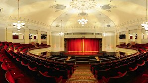 Un vaste auditorium circulaire doté de gradins, une scène centrale surmontée d’un rideau rouge et un plafond richement décoré.