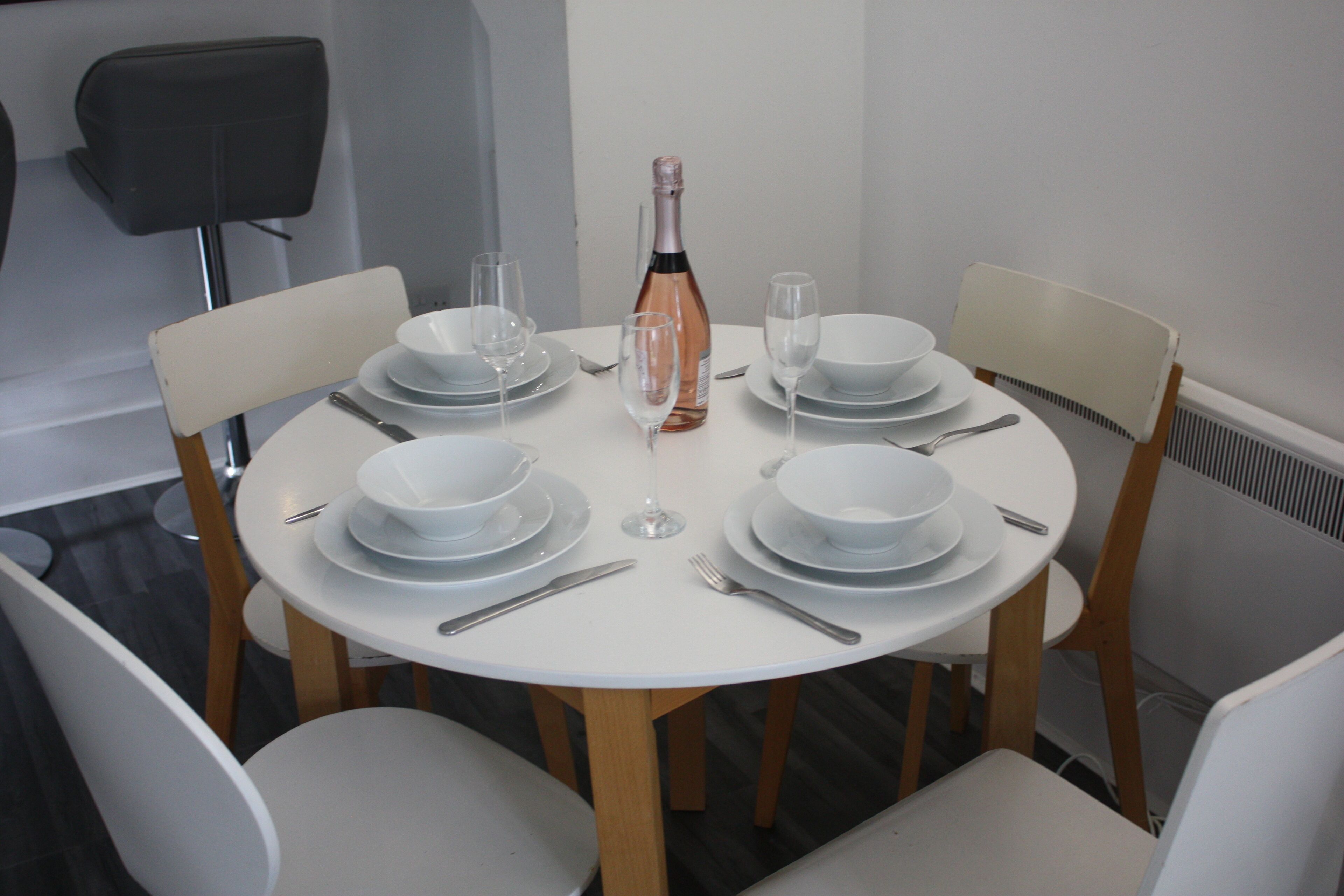 Une table de salle à manger ronde, avec de l’assiette blanche, des couverts et une bouteille de vin, entourée de chaises blanches.