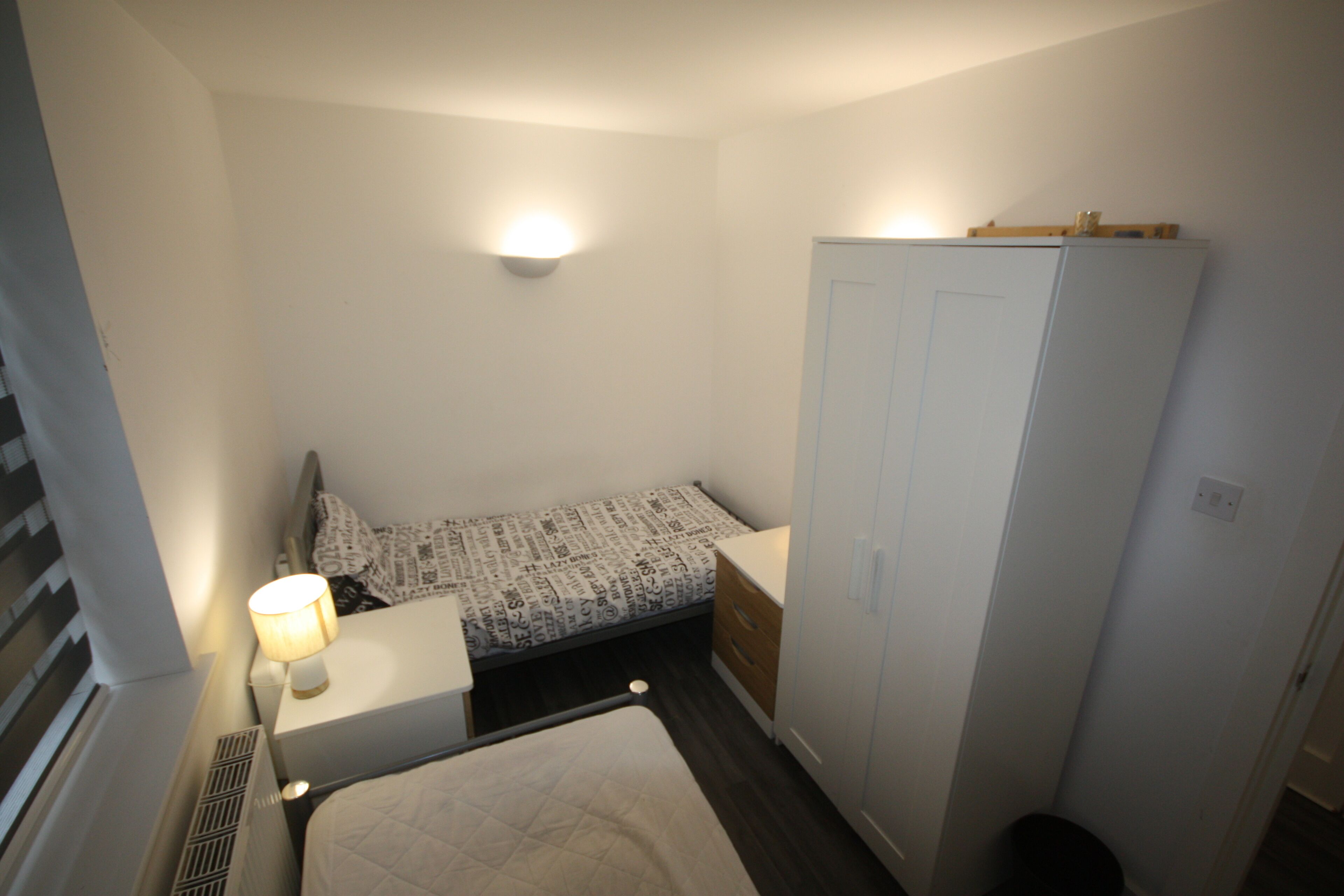 A small, compact bedroom with a single bed, a nightstand with a lamp, a wardrobe, and a wall-mounted light fixture.
