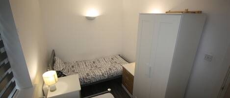 A small, compact bedroom with a single bed, a nightstand with a lamp, a wardrobe, and a wall-mounted light fixture.