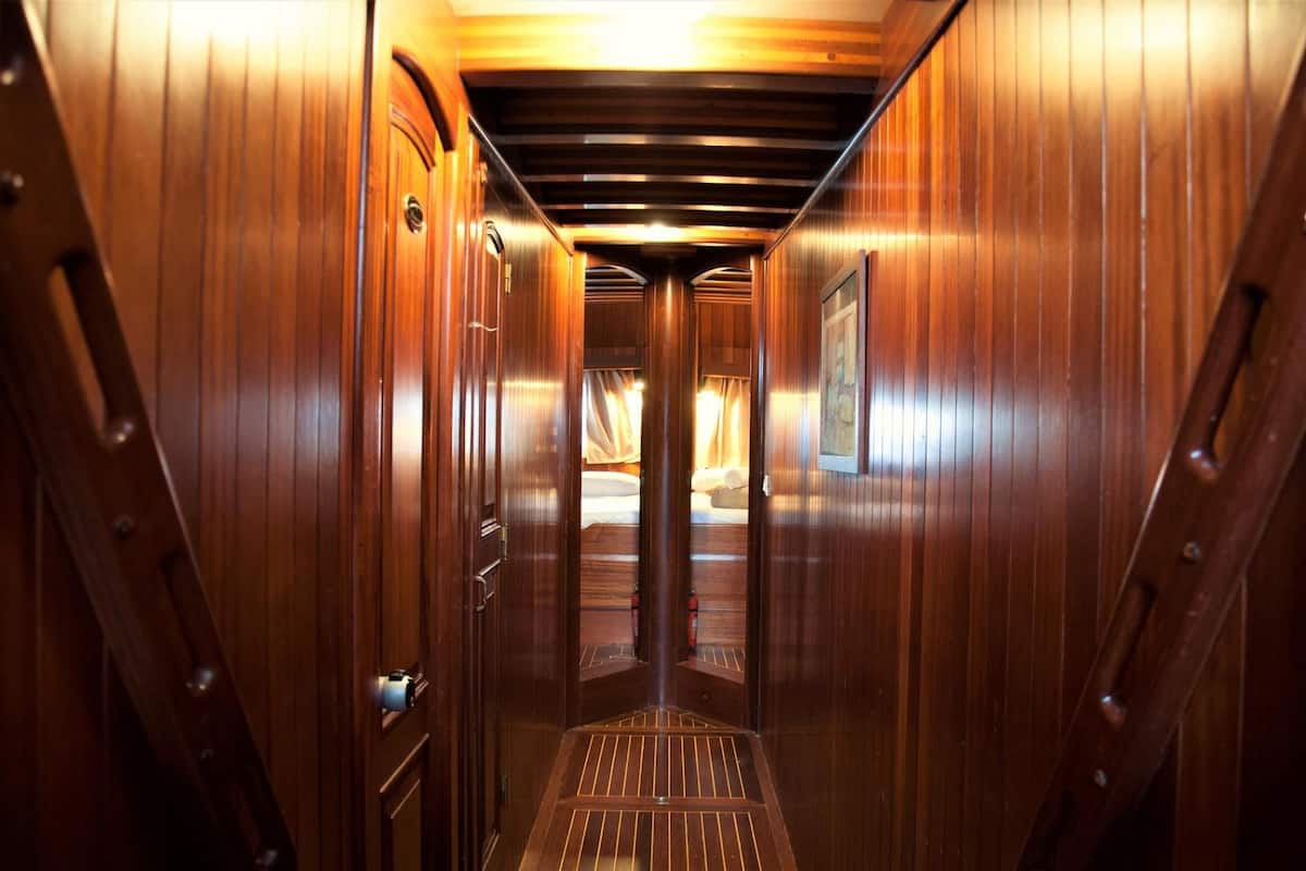 A narrow, wooden corridor with a metal door at the end leading to a room with a bed.