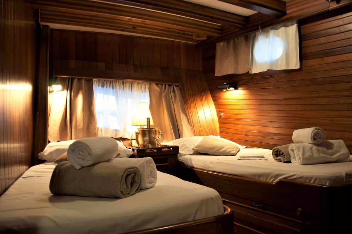 A cabin-style room with wooden walls and ceiling, two beds with white linens, and a nightstand with a lamp and a glass.