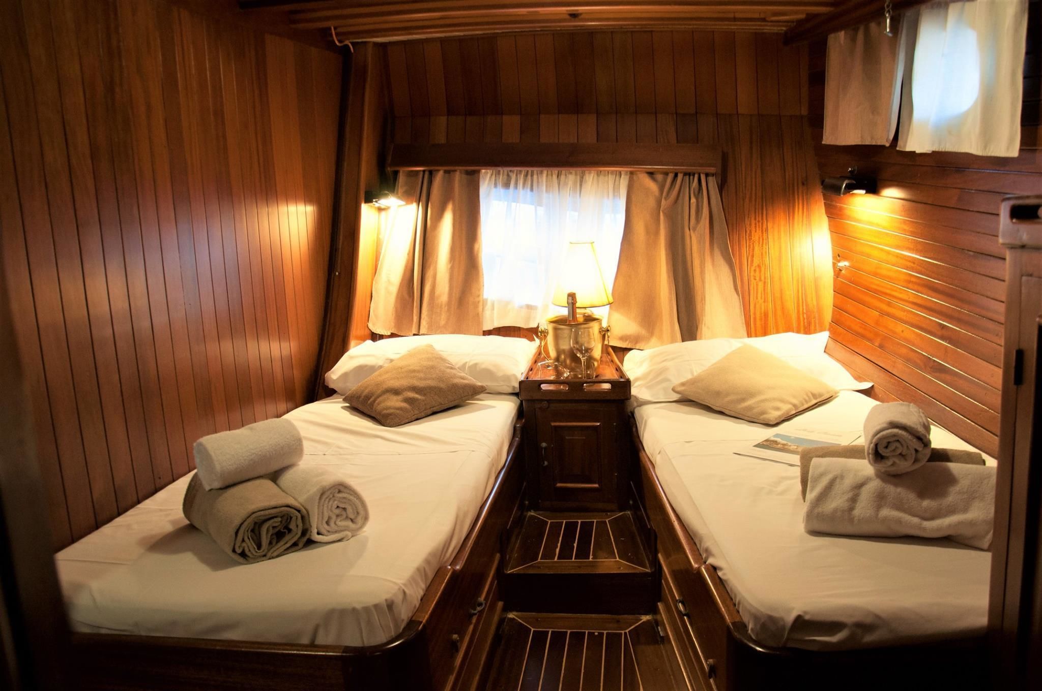 A cabin with two single beds, wooden walls, a small table with a lamp, and a window with curtains.