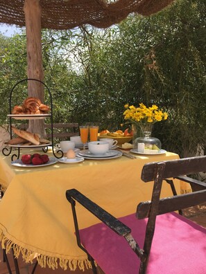 Un montaje para comer al aire libre con mantel amarillo, una silla de mimbre y una variedad de alimentos para el desayuno, como pan, frutas y bebidas.