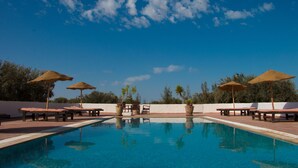 A pool area with lounge chairs and umbrellas, surrounded by a white wall and trees. - Domaine El-Manar (Zarzis)