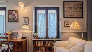 A cozy room with a wooden desk, a bookshelf, a white sofa, and a window with curtains. - La Curandera (Rosta)