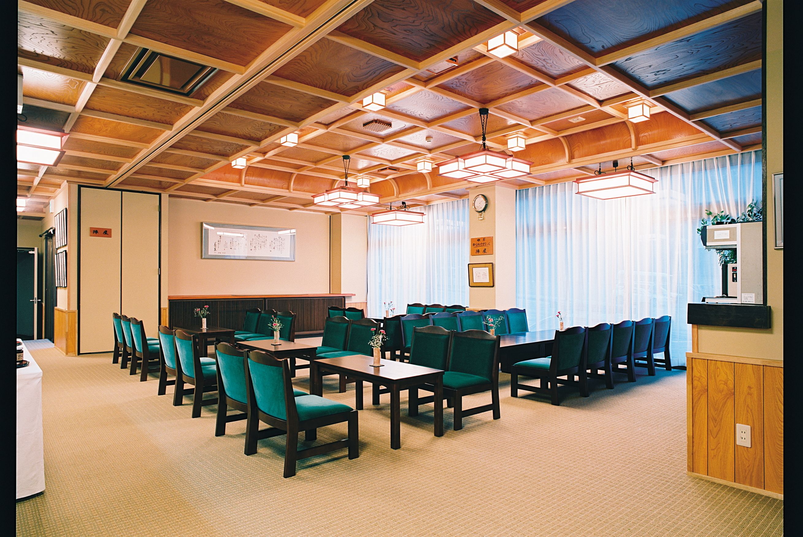 A conference room with wooden ceiling beams, green chairs, and tables. There are windows with curtains and a clock on the wall.
