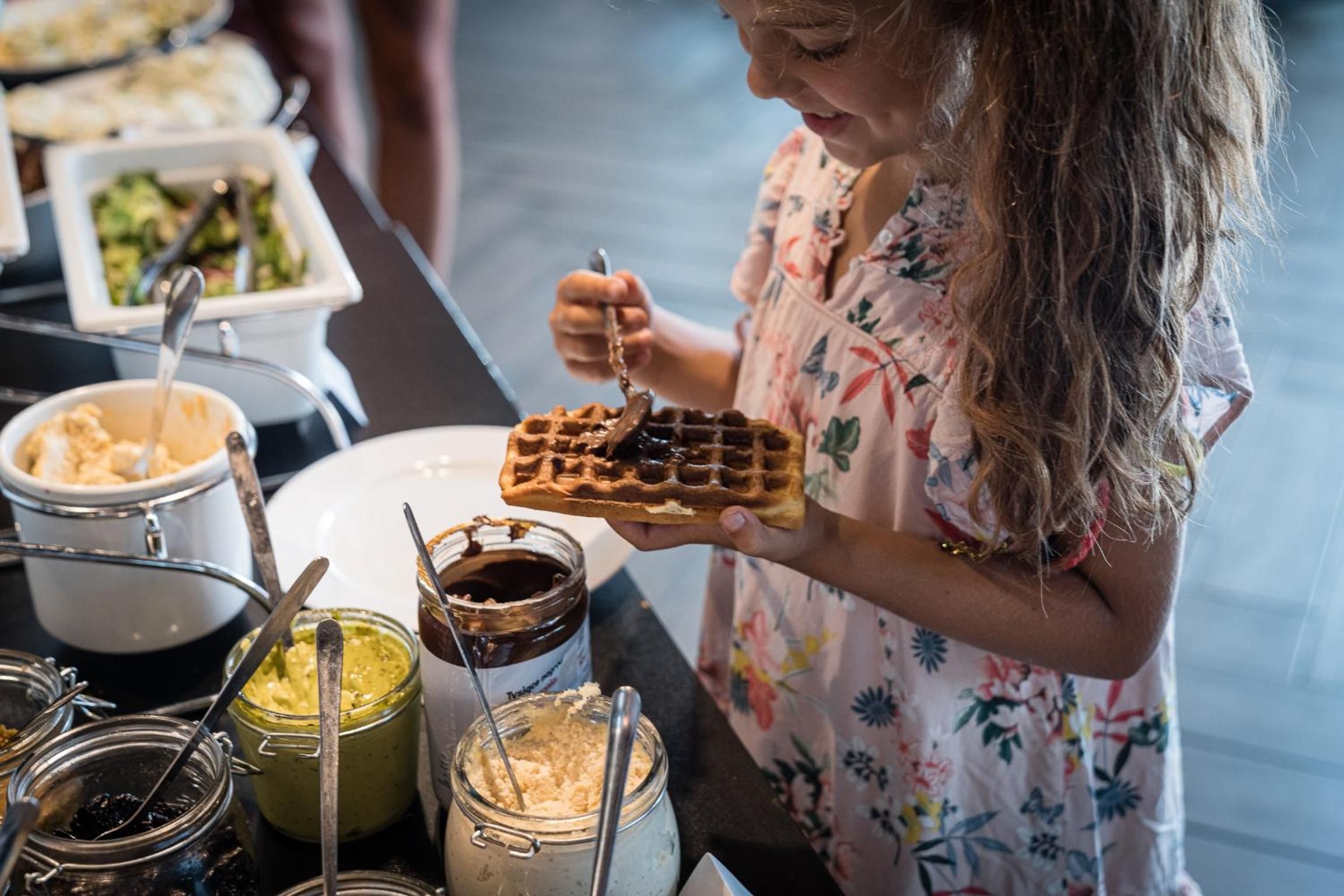 A person holding a waffle with syrup, with various food items and utensils in the background.