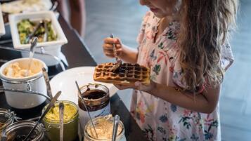 A person holding a waffle with syrup, with various food items and utensils in the background.
