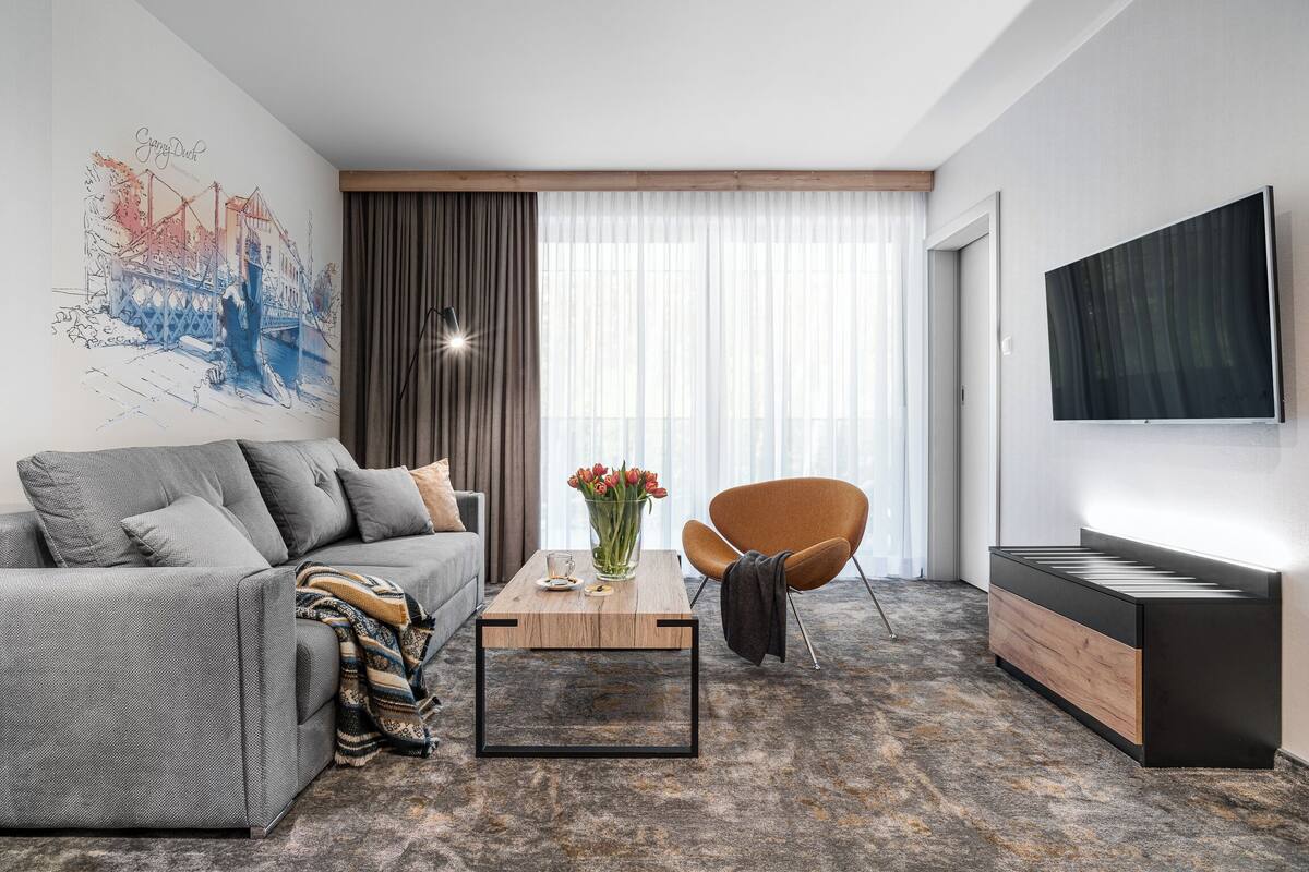A modern living room with a grey sofa, a wooden coffee table, a vase of flowers, a brown armchair, and a TV on the wall.