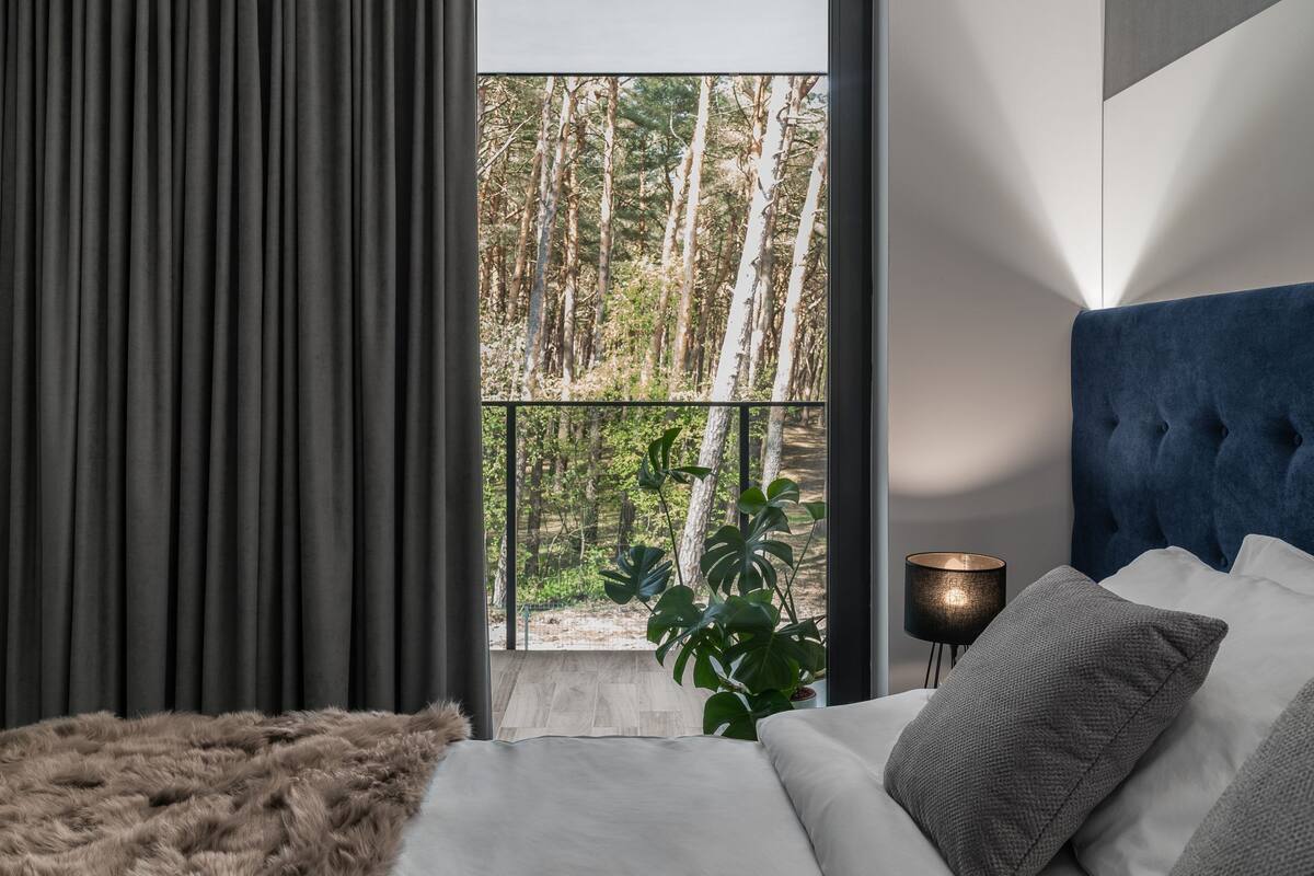 A bedroom with a bed, a view of trees, a plant, and a lamp.