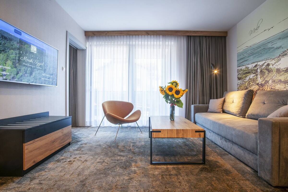 A modern living room with a sofa, a coffee table, a TV mounted on the wall, and a large window with curtains.