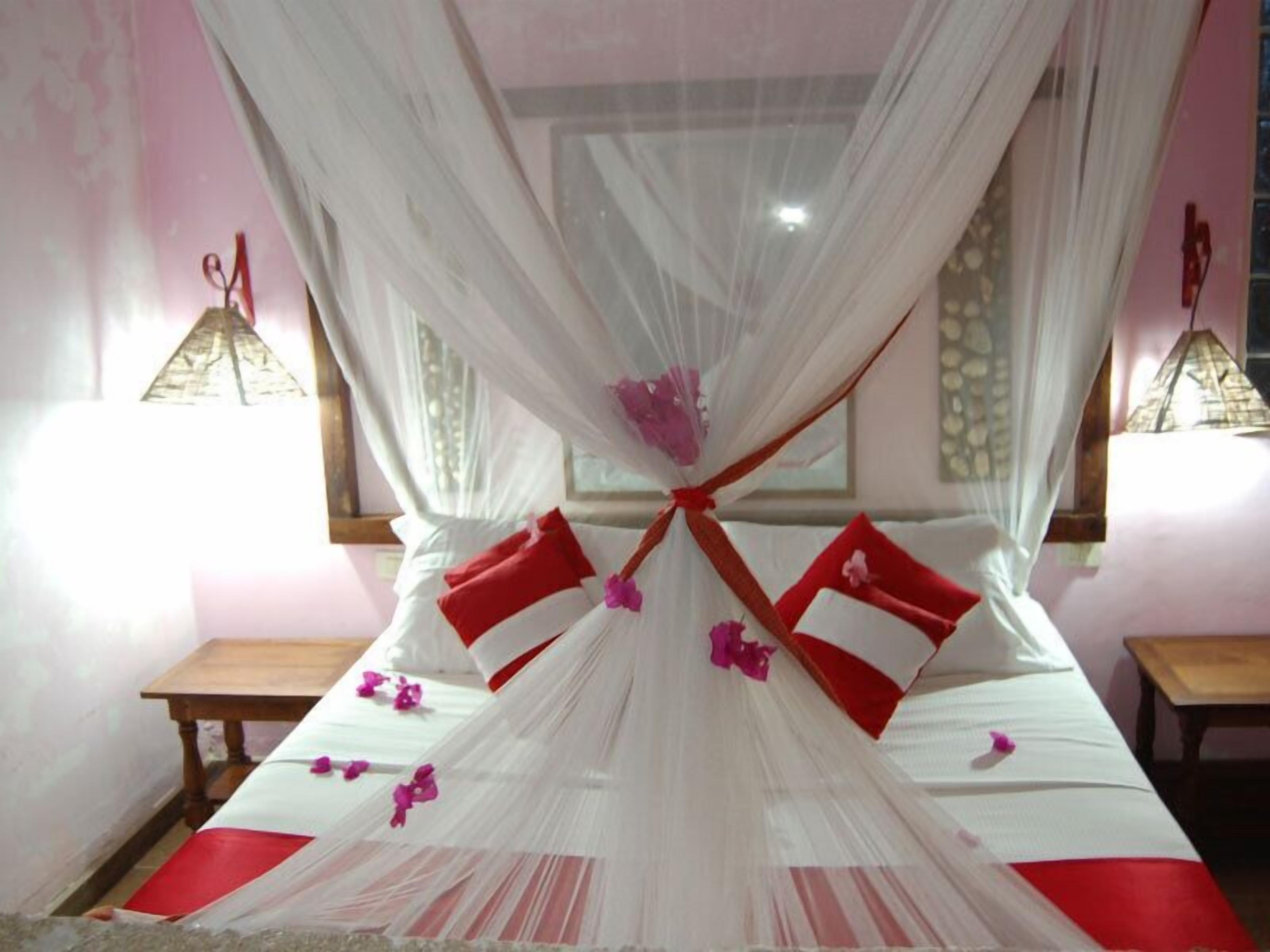 Una cama con dosel, almohadas rojas y una colcha roja y blanca. Hay dos mesitas de noche y una ventana con cortinas.