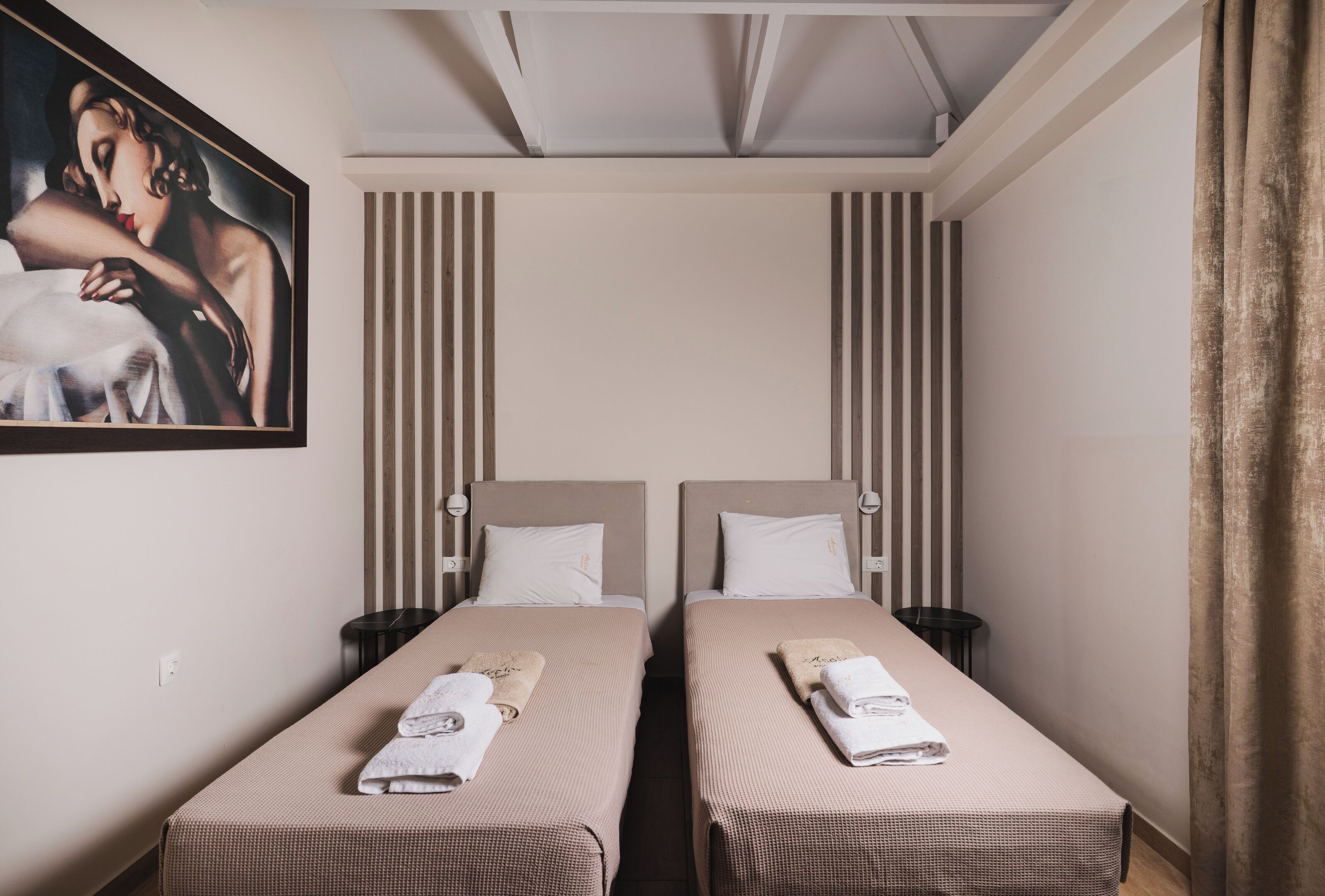 Camera d'albergo con due letti singoli, ognuno con un cuscino e un asciugamano. Sul muro c'è un quadro e una finestra con tende.