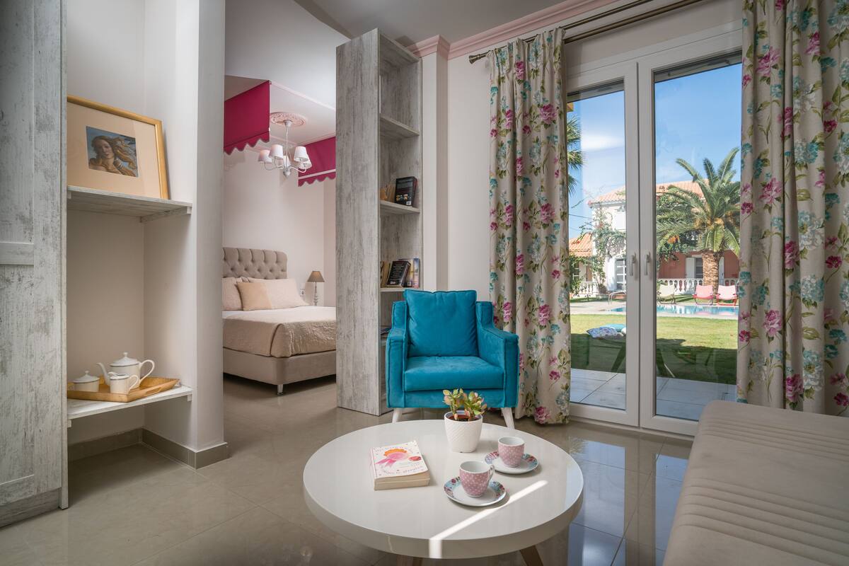 Un soggiorno moderno con una poltrona blu, un tavolino rotondo con un libro e un teiera, e vista su una piscina e palme attraverso ampie finestre.