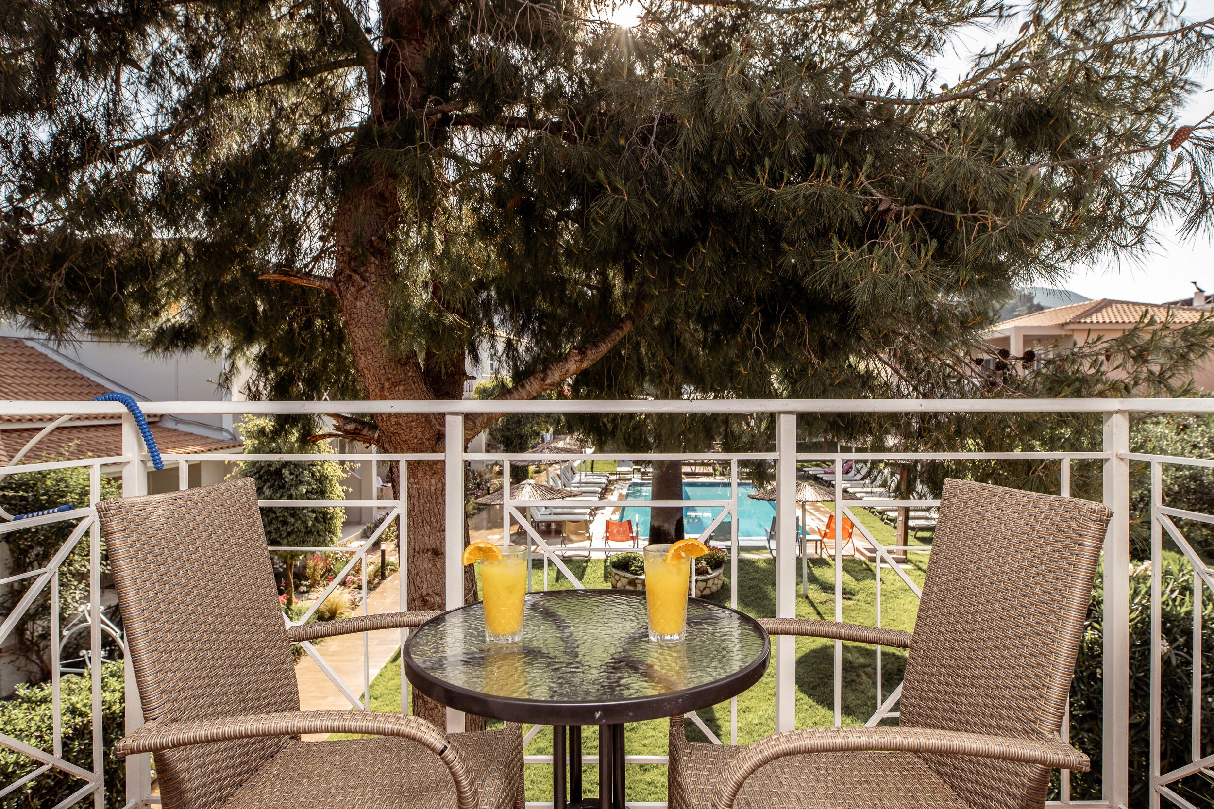 Een balkon met rieten stoelen en een tafel, twee drankjes, en een zwembad op de achtergrond.