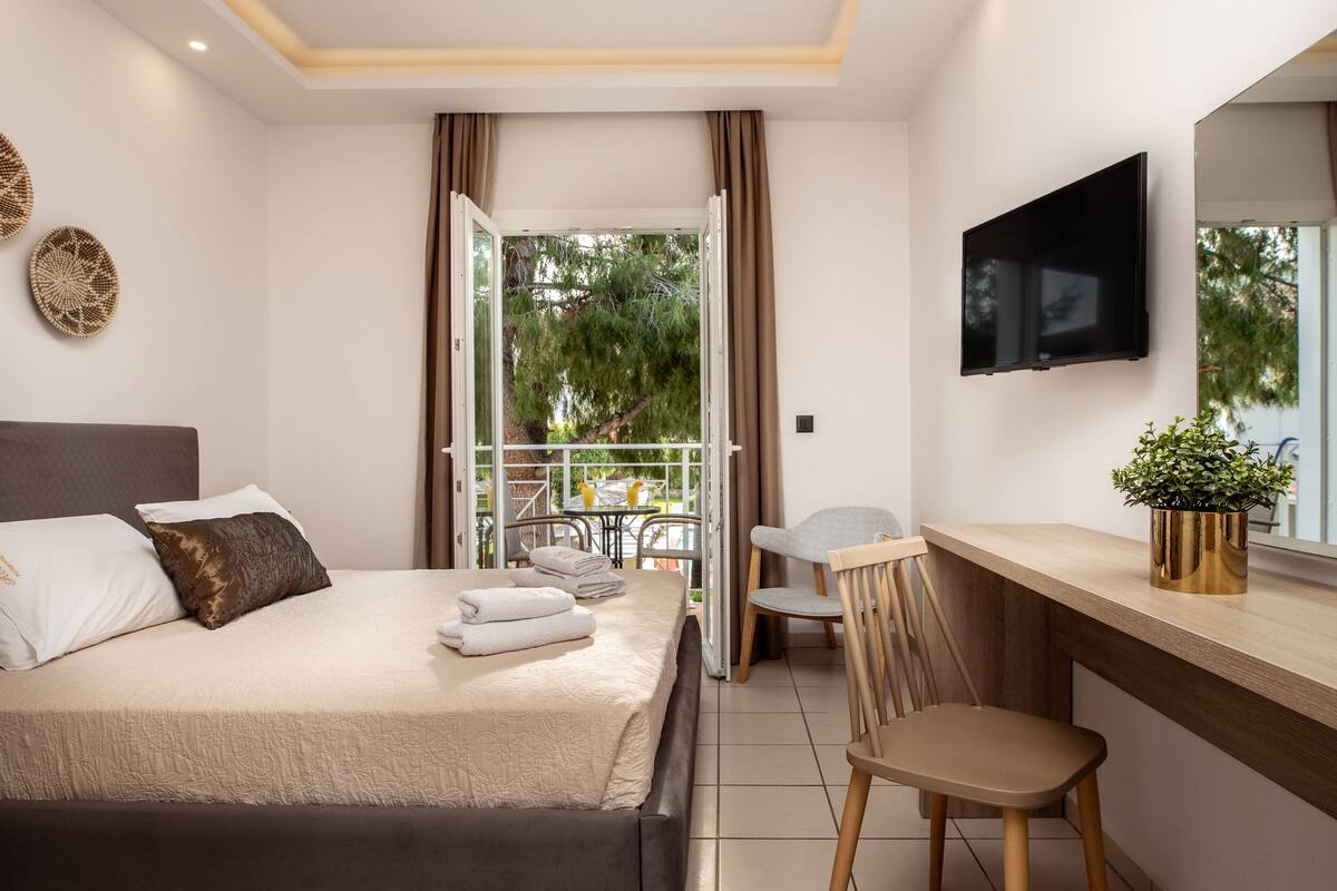 Una camera d'albergo moderna con un letto, una scrivania, una sedia, una TV e un balcone con vista sugli alberi.