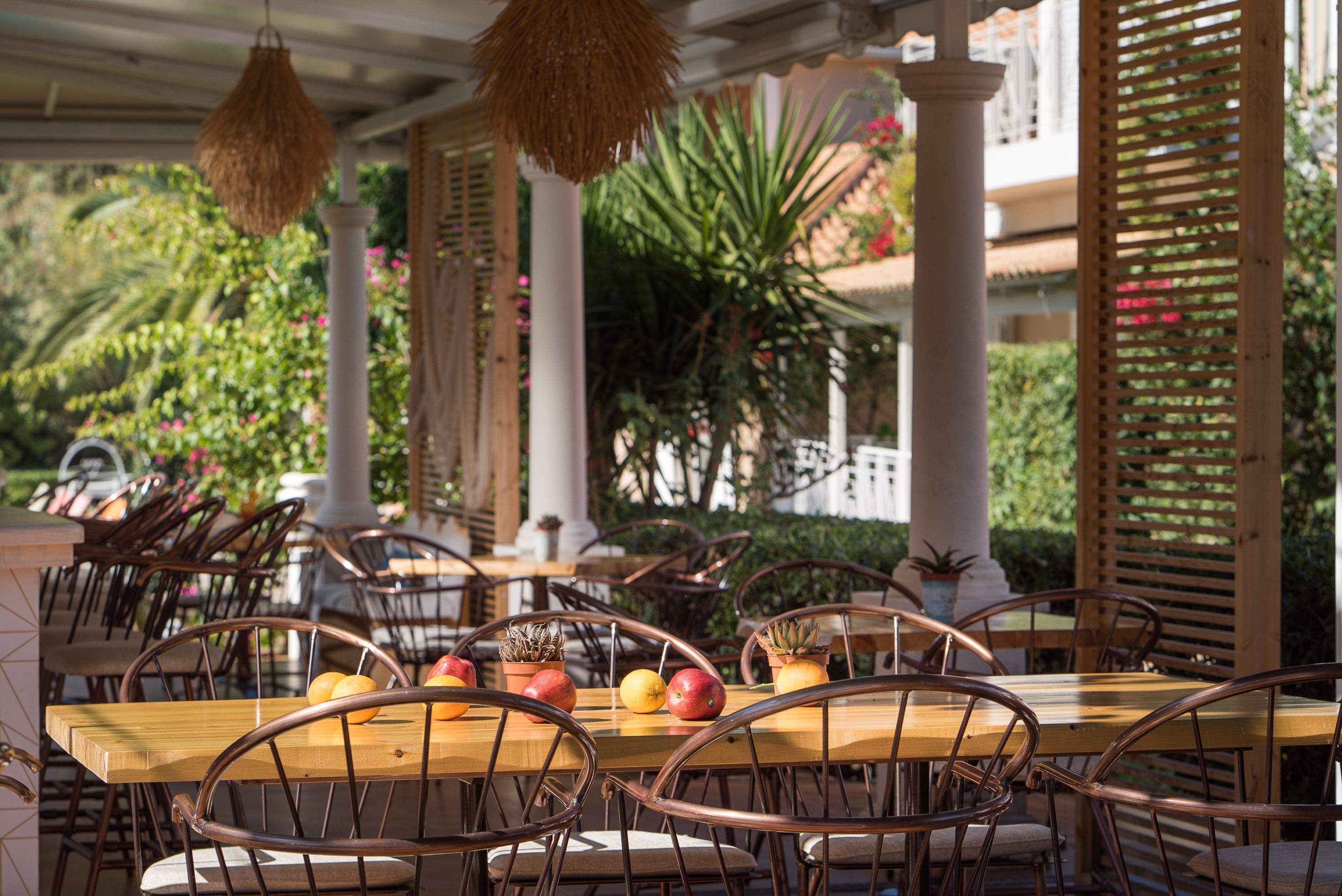 An outdoor dining area with wooden tables and chairs, surrounded by greenery and columns.
