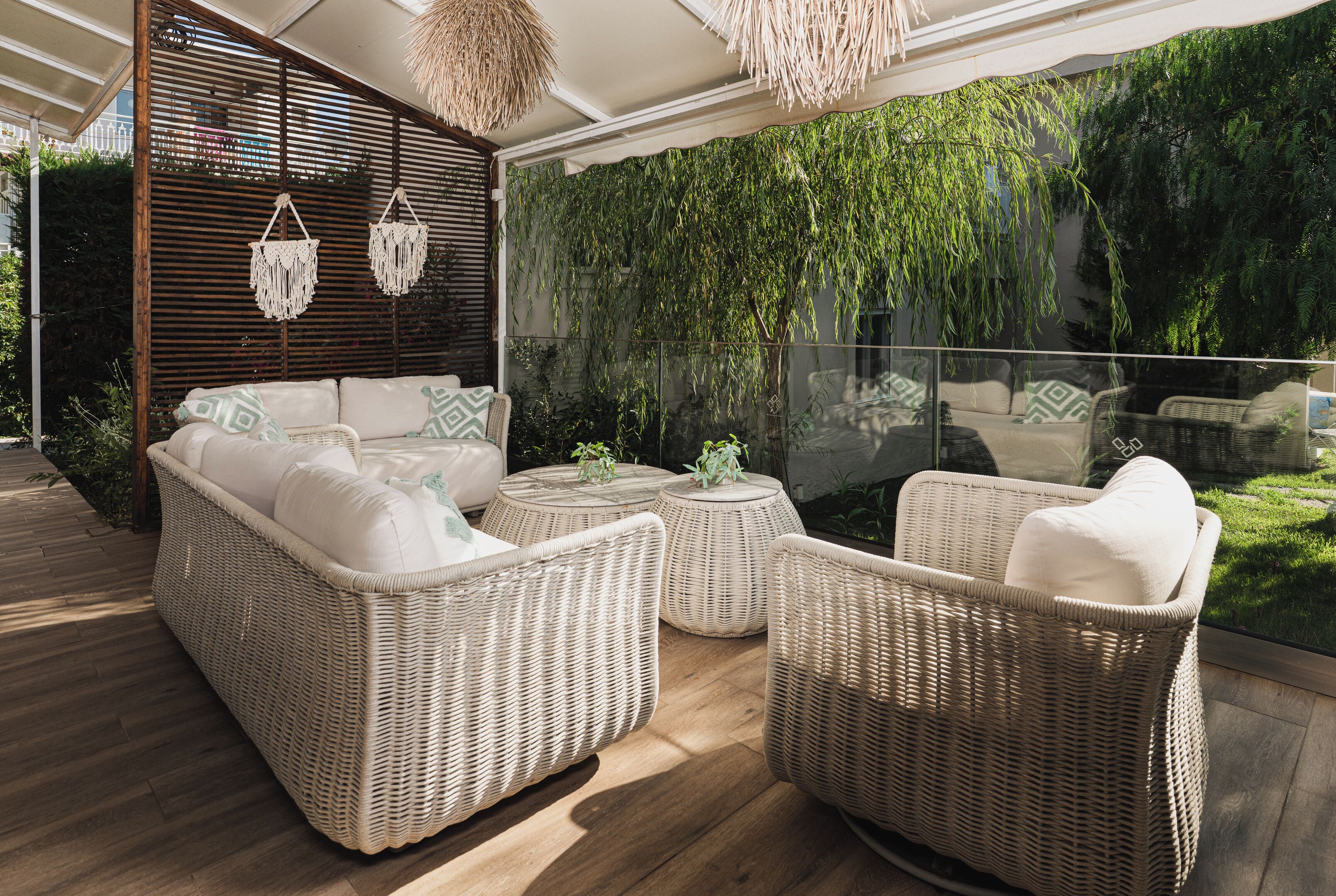 A wicker sofa set with cushions, a round table, and hanging macramé light fixtures in a sunlit outdoor area with a view of greenery.