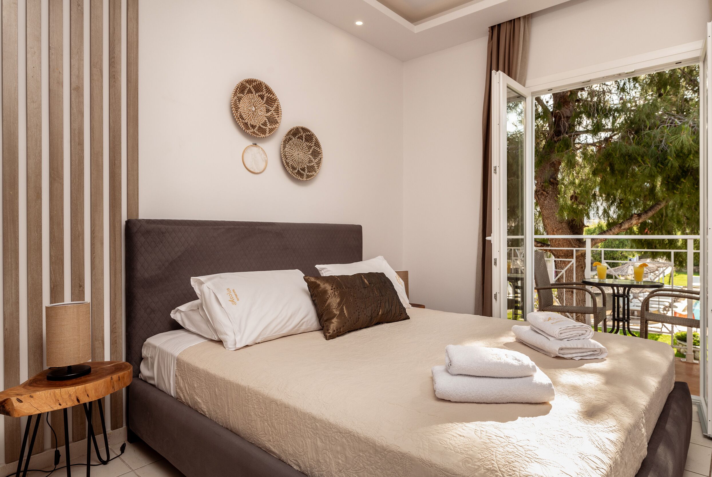 A bedroom with a large bed, a wooden side table, and a balcony with outdoor furniture.