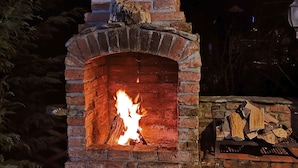 A brick-built outdoor fireplace with a fire burning inside, surrounded by logs and a metal bucket. - Casa Crina (Saliste)