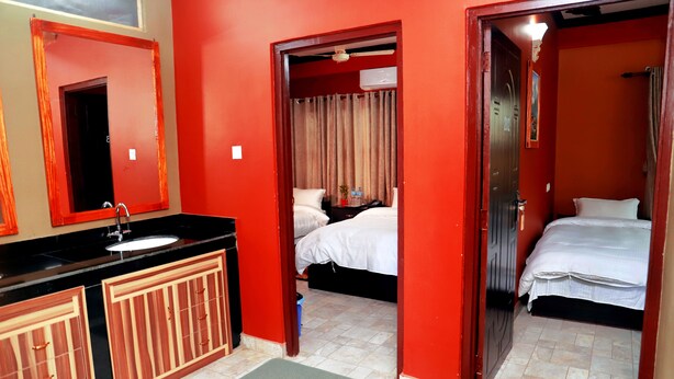 Een hotelkamer met een rode muur, een bed, een nachtkastje, een spiegel en een badkamer met een wastafel en kastjes.
