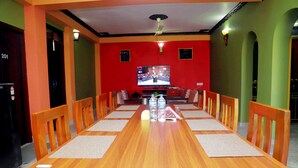 Een lange houten eettafel met stoelen, een televisiescherm en een plafondventilator in een kamer met groene muren en een zwarte deur.