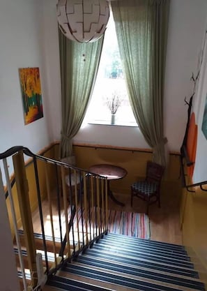 Staircase leads to an upper floor, flanked by a window with sheer curtains and a small table for relaxation.