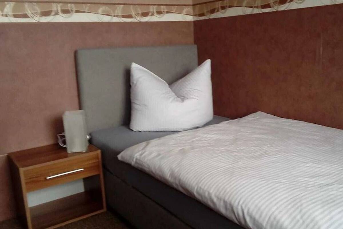 Una cama individual con cabecera gris y almohada blanca, una mesita de noche de madera con cajón, y una pared lisa con un detalle decorativo.