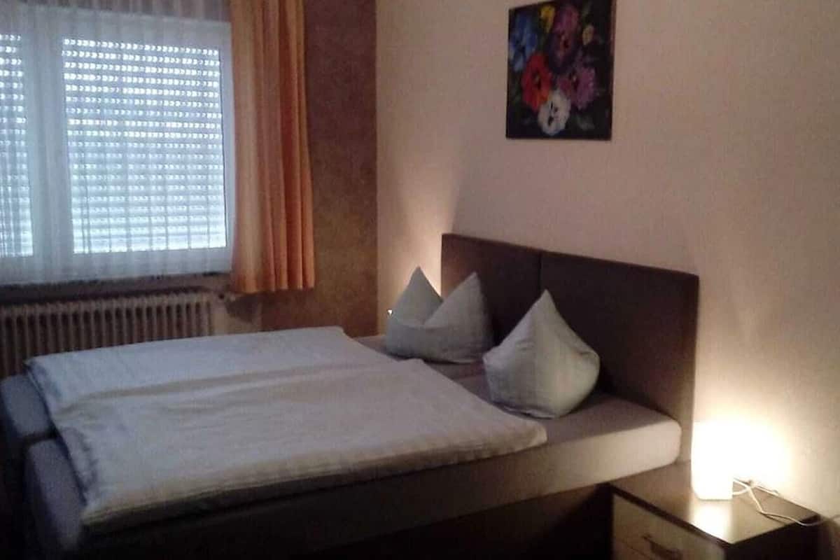 Un dormitorio con cama, almohadas, mesita de noche, lámpara, radiador y ventana con cortinas.