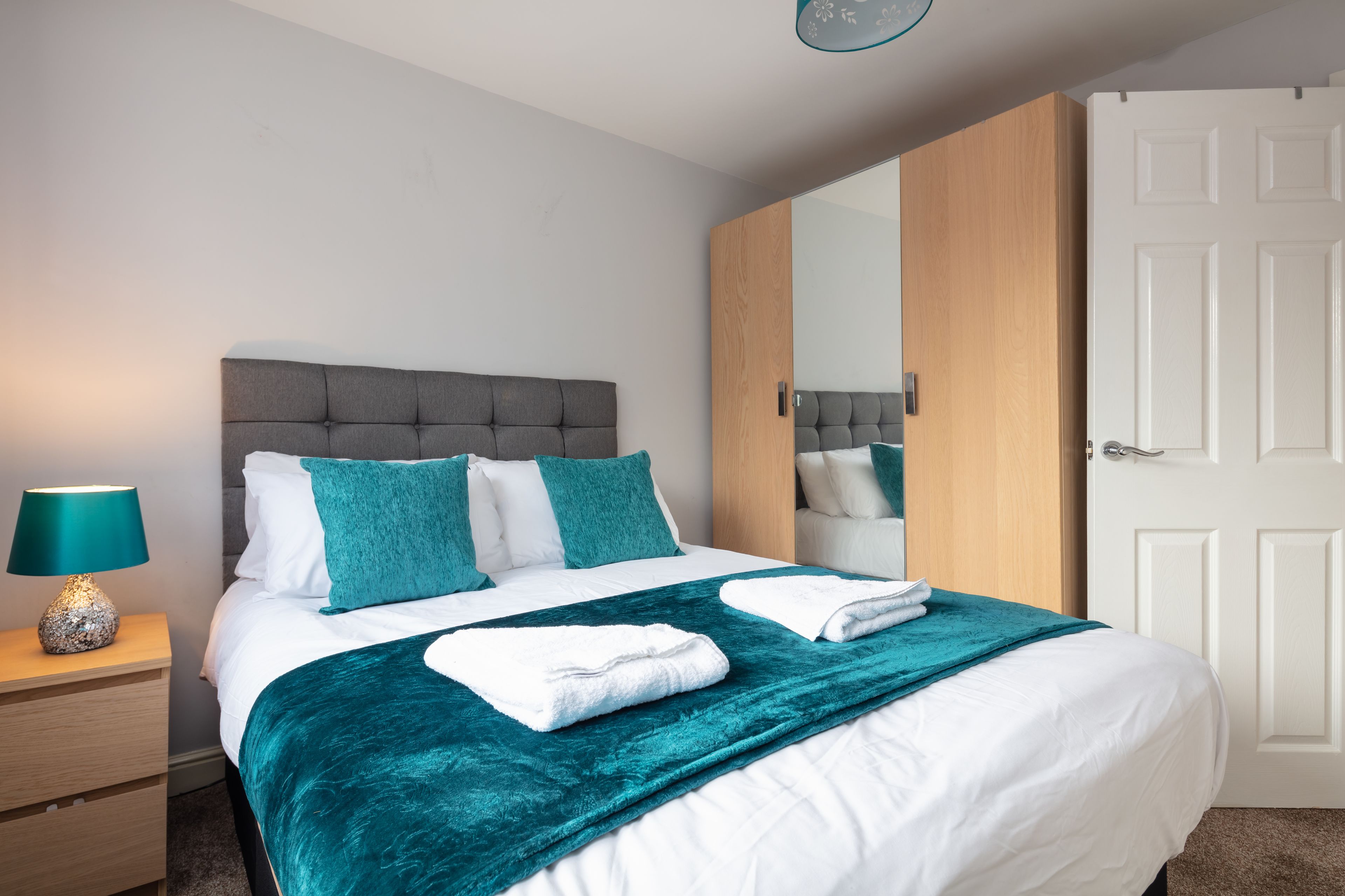 A neatly made bed with a teal throw blanket, a bedside lamp, and a wooden nightstand.