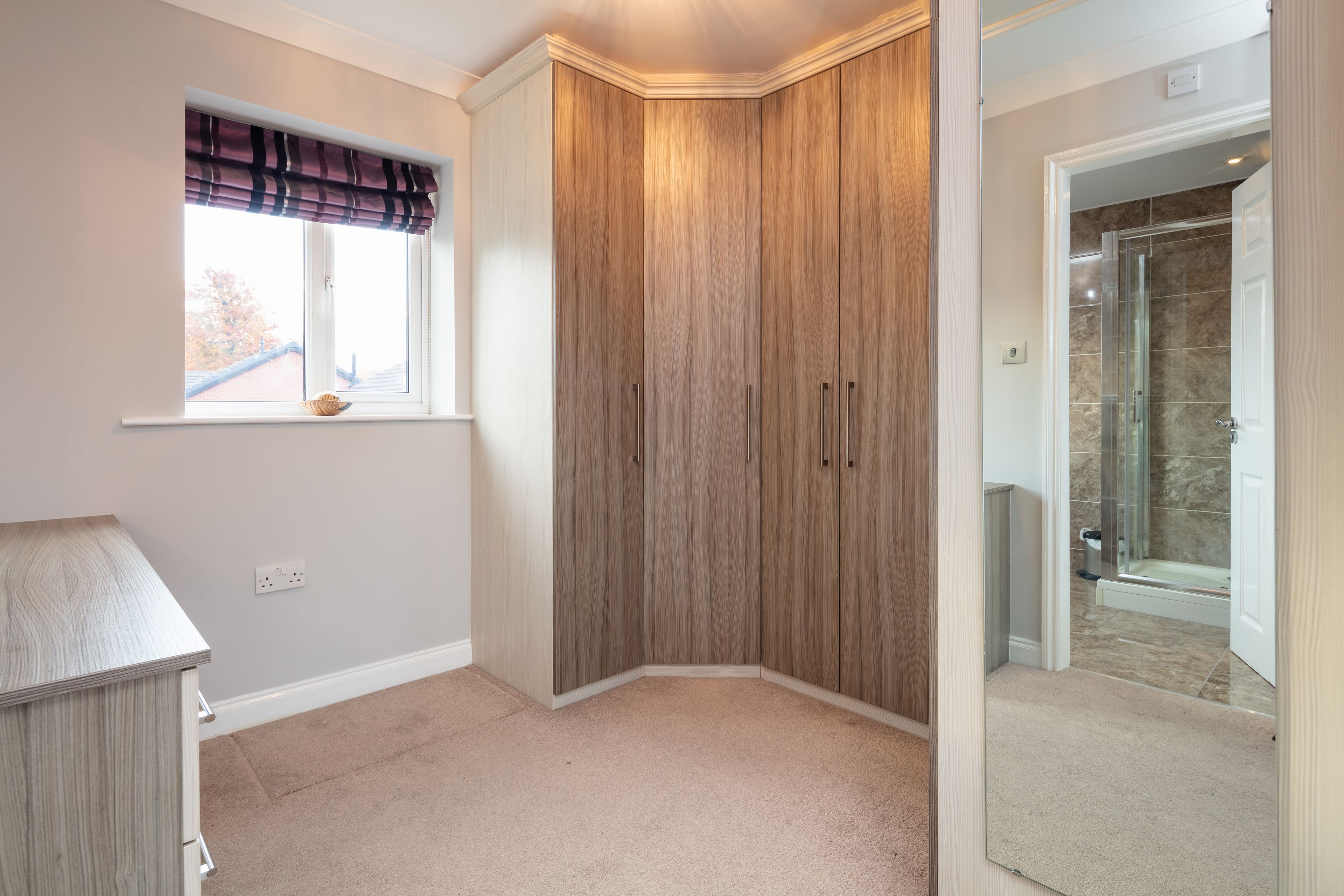 A corner bedroom with a large wardrobe, a window with a plaid valance, and a view into a bathroom with a glass shower.