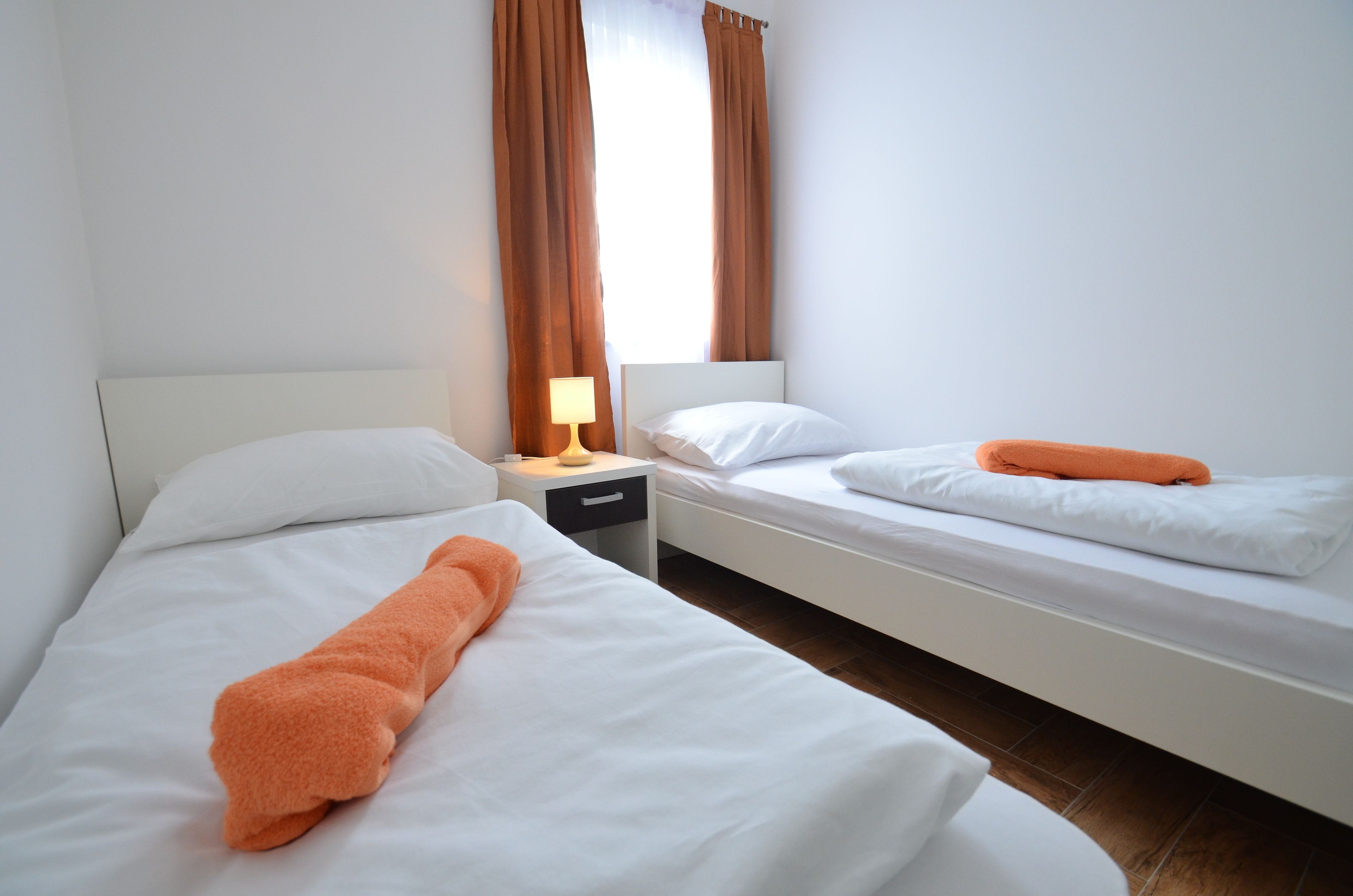 A hotel room with two single beds, a nightstand with a lamp, a window with curtains, and a wooden floor.