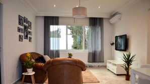 A modern living room with a brown leather sofa, a white coffee table, a TV, and a large window with curtains. - Cosy S3 At Sidi Bou Said Village (Sidi Bou Said)