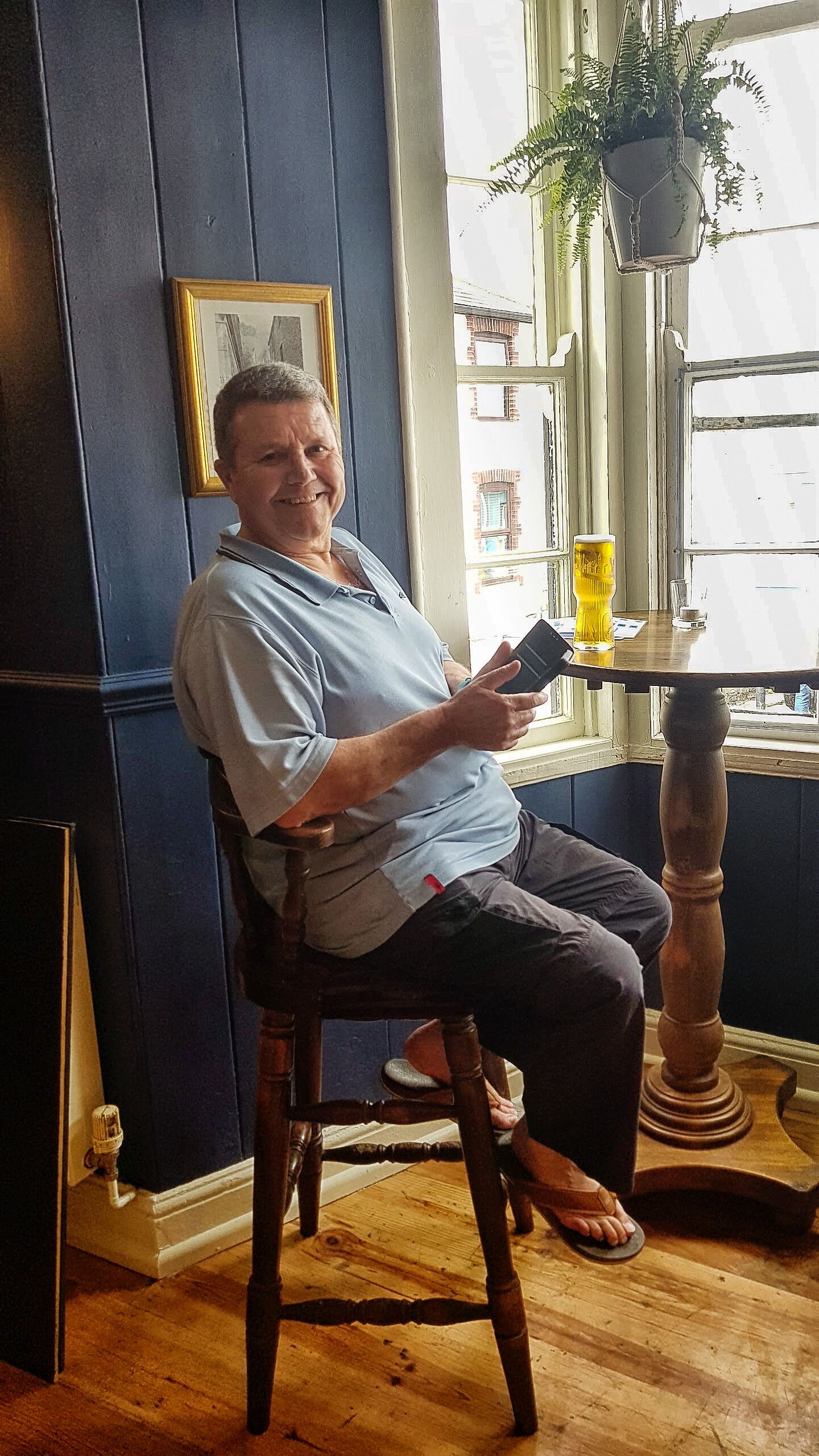 A person seated on a wooden chair in a room with dark blue walls, a window with a view of buildings, and a table with a yellow object on it.