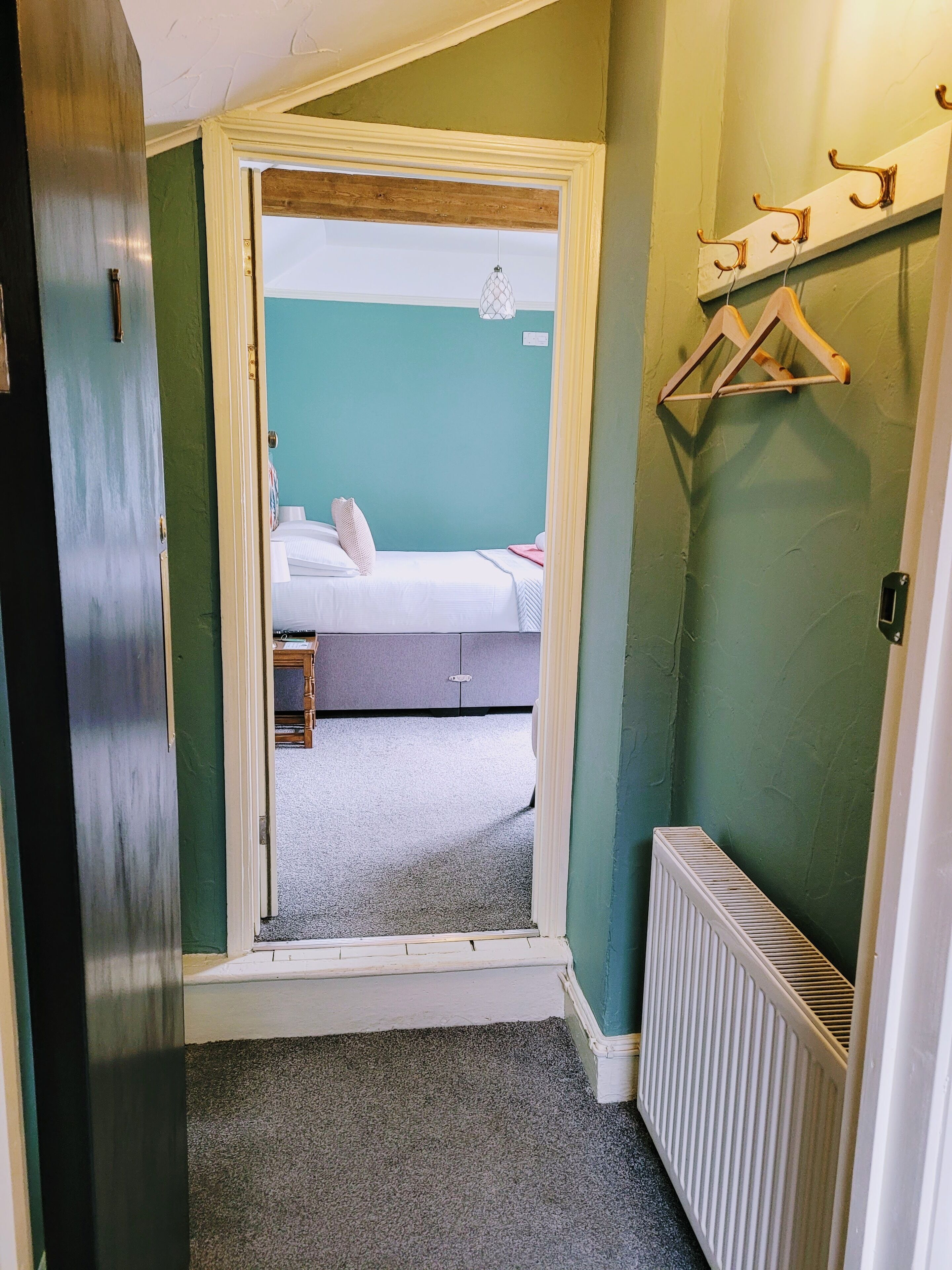 A narrow hallway leading to a bedroom with a bed, a radiator, and a coat rack.