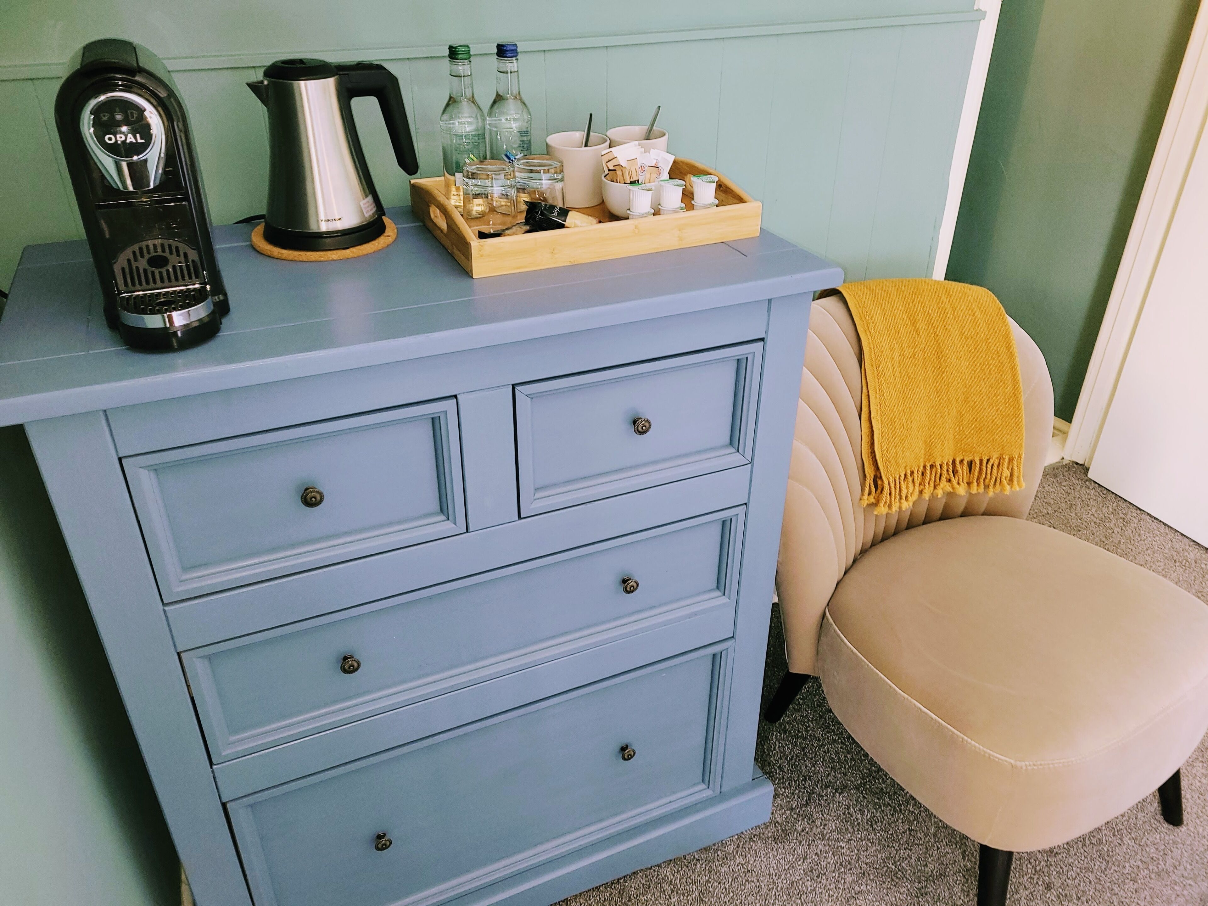 A light blue dresser with drawers, a coffee maker, a kettle, and a tray with bottles and cups.