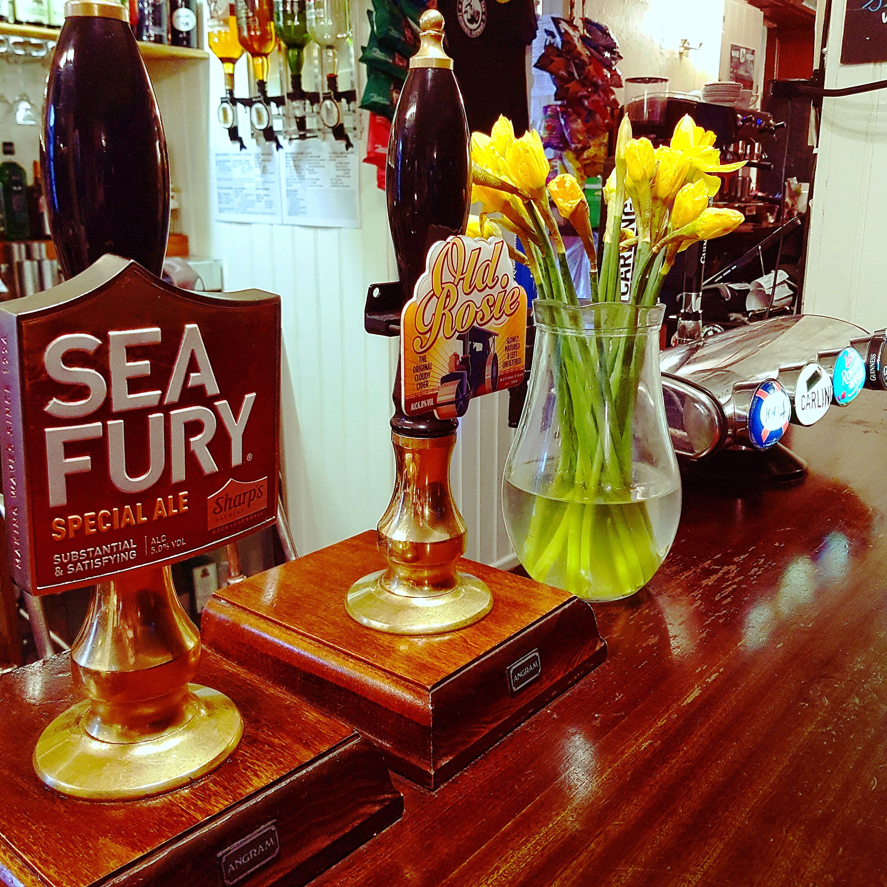 Um balcão de bar com um letreiro 'Sea Fury Special Ale', uma garrafa de cidra 'Old Rosie' e um vaso com flores amarelas.