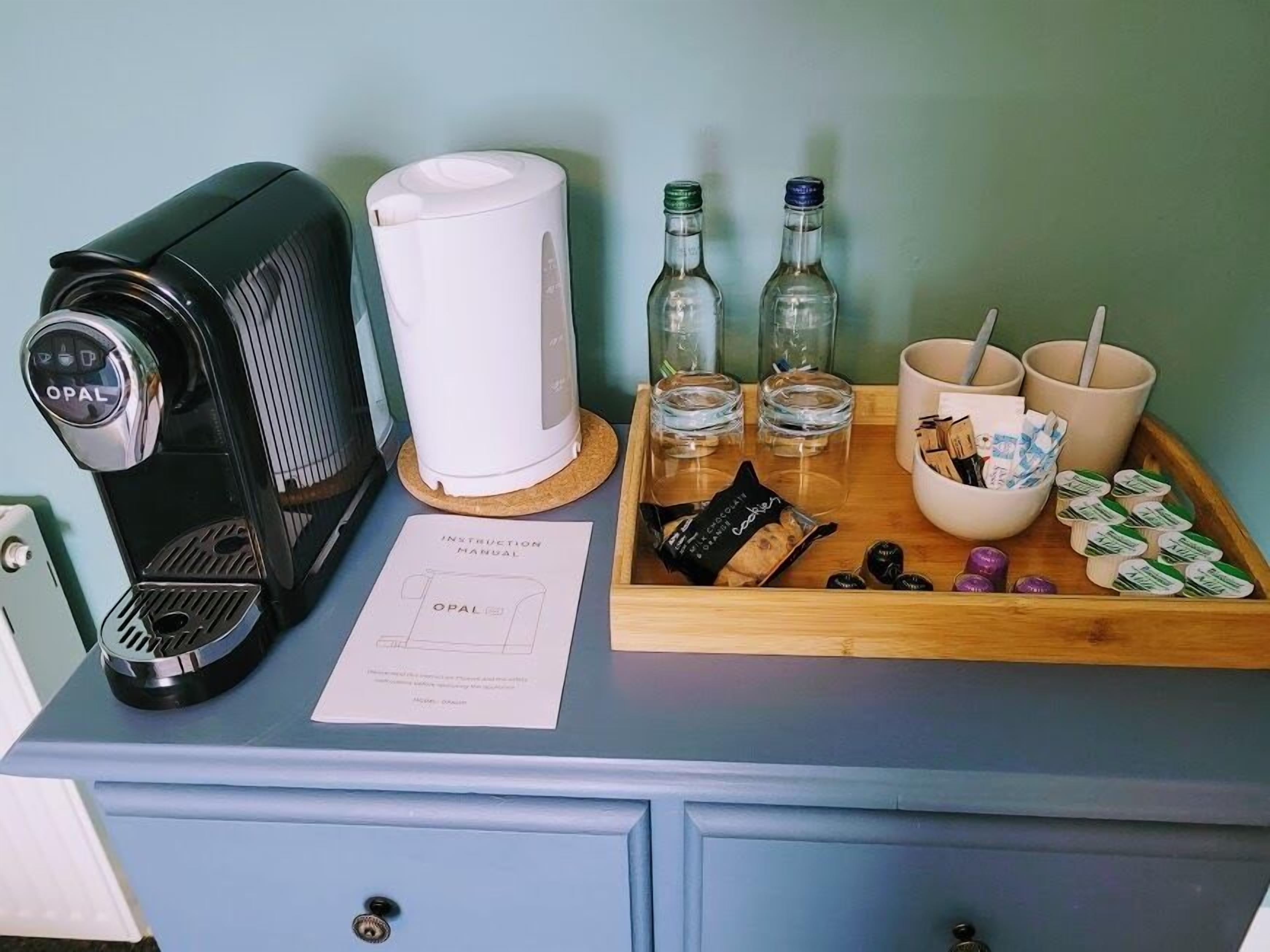 A coffee maker, a white electric kettle, two bottles, two glasses, a tray with cookies, and a bowl with straws and snacks.