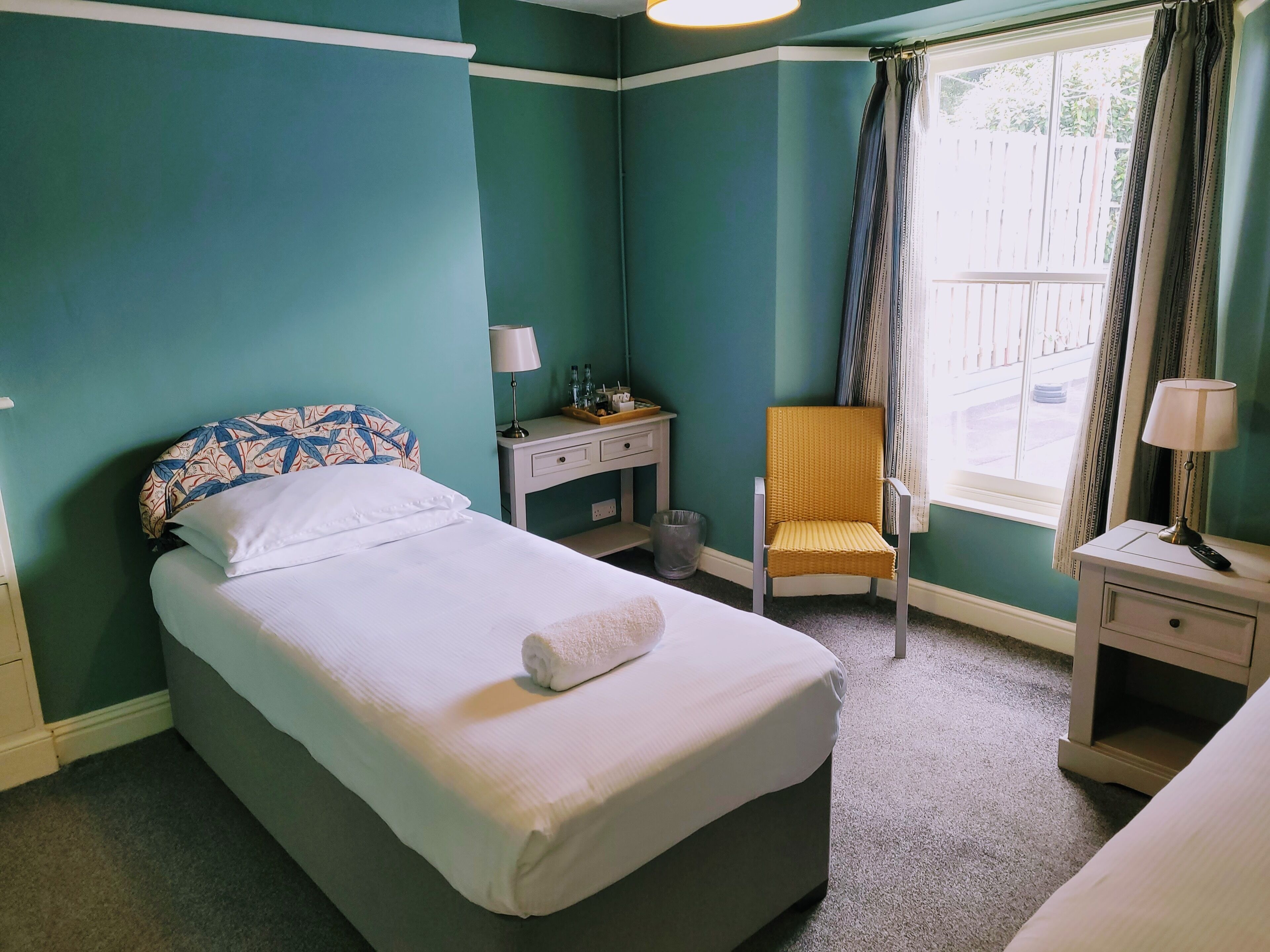A neatly made bed with a white comforter and a rolled towel, a small bedside table with a lamp, a chair with a cushion, and a window with curtains.