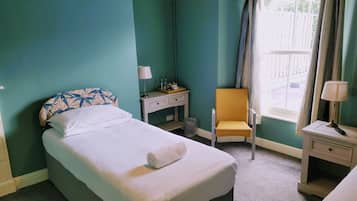 A neatly made bed with a white comforter and a rolled towel, a small bedside table with a lamp, a chair with a cushion, and a window with curtains.